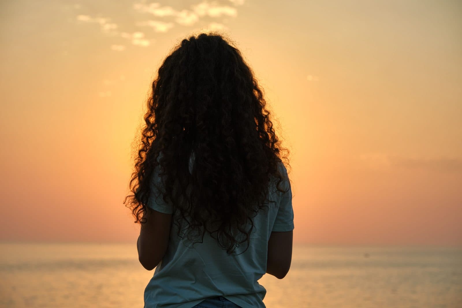 Mulher de cabelos cacheados, de costas, contempla o mar sob luz dourada