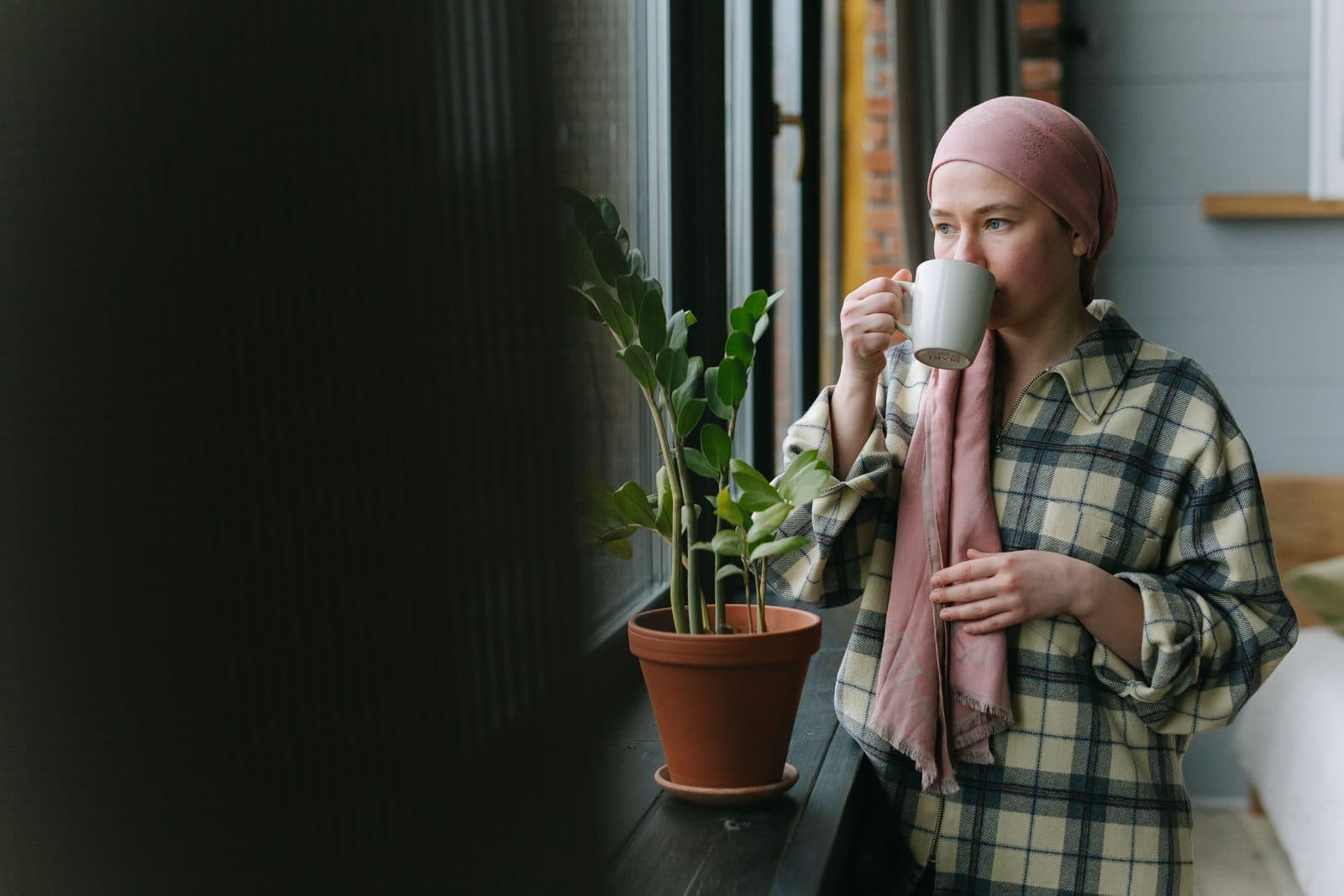 Mulher de lenço na cabeça toma café serena junto à janela com planta